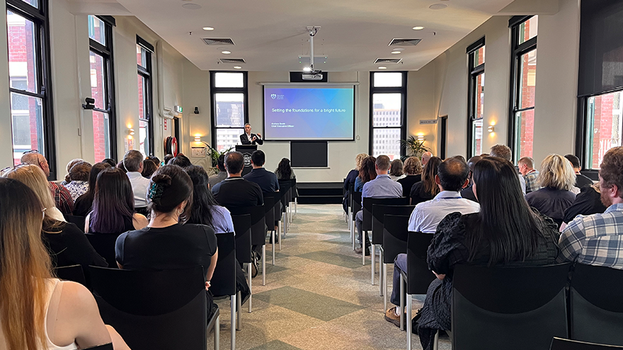A speaker presents to a seated audience in a conference room, with a presentation slide titled "Setting the foundations for a bright future" displayed on a screen at the front.
