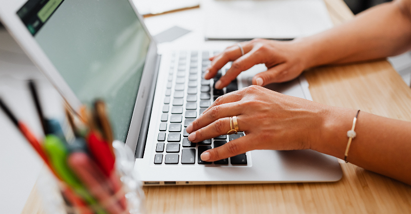 Two hands typing on a laptop at a desk. A container of pens is blurred in the foreground.