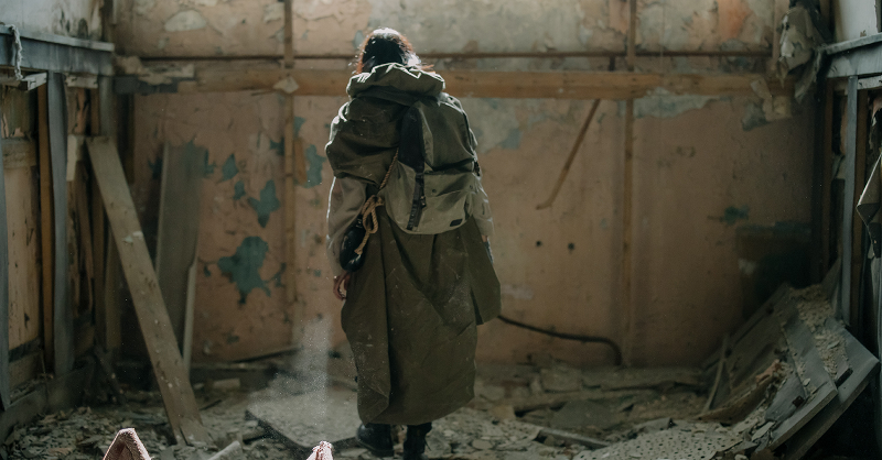 A figure dressed in a big jacket and backpack stands in a dystopian room that has been destroyed over time, their back is to the camera.