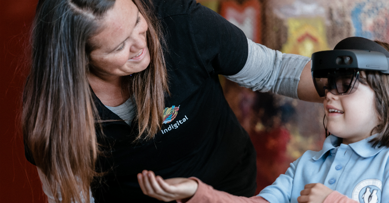 Child uses an AR device, holding out her hand while an educator offers support.