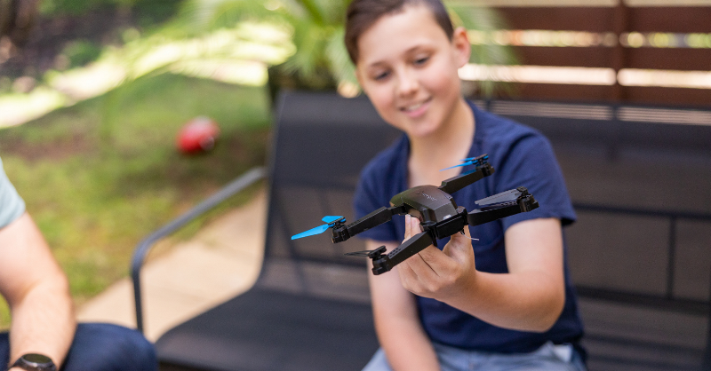 A young boy sitting outside on an outdoor lounge, holding a black and blue drone in his hand, inspecting it and smiling. 